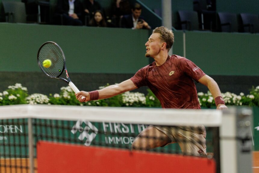 Male tennis player in a maroon shirt lunges to hit a yellow ball with a racket on an indoor court, spectators in the stands behind him.