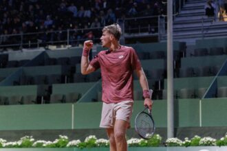 Tennis player in maroon shirt raises fist in celebration on a court with spectators in the stands behind him.