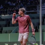 Tennis player in maroon shirt raises fist in celebration on a court with spectators in the stands behind him.