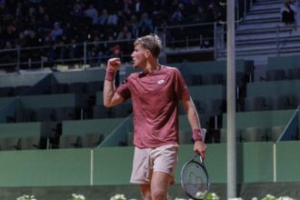 Tennis player in maroon shirt raises fist in celebration on a court with spectators in the stands behind him.