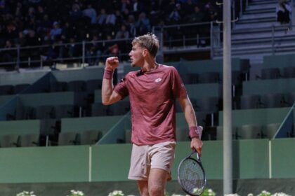 Tennis player in maroon shirt raises fist in celebration on a court with spectators in the stands behind him.