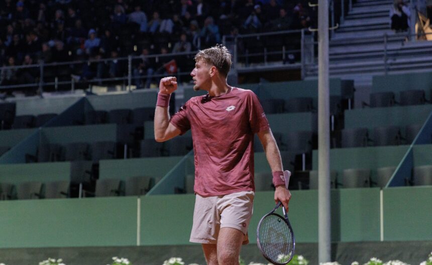 Tennis player in maroon shirt raises fist in celebration on a court with spectators in the stands behind him.