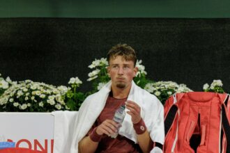 Male athlete resting with a white towel over his shoulders, holding a water bottle, red backpack beside him with a backdrop of white daisies.