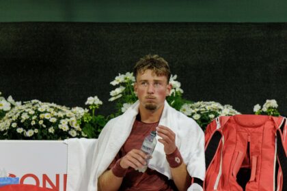 Male athlete resting with a white towel over his shoulders, holding a water bottle, red backpack beside him with a backdrop of white daisies.