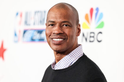 Portrait of a smiling Black man with a shaved head, wearing a checkered shirt and dark sweater at a media event with NBC branding in the background.