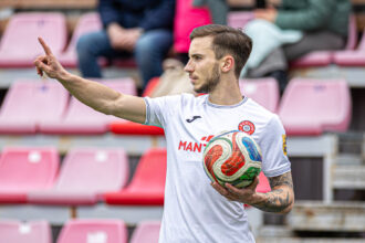 Soccer player in a white jersey holding a multicolor ball and pointing with his left arm in a stadium with pink seats.
