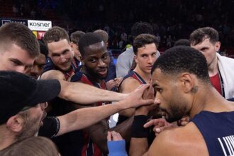 Basketball players in navy and red uniforms form a huddle with hands joined in the center before play in a crowded arena.