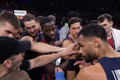 Basketball players in navy and red uniforms form a huddle with hands joined in the center before play in a crowded arena.