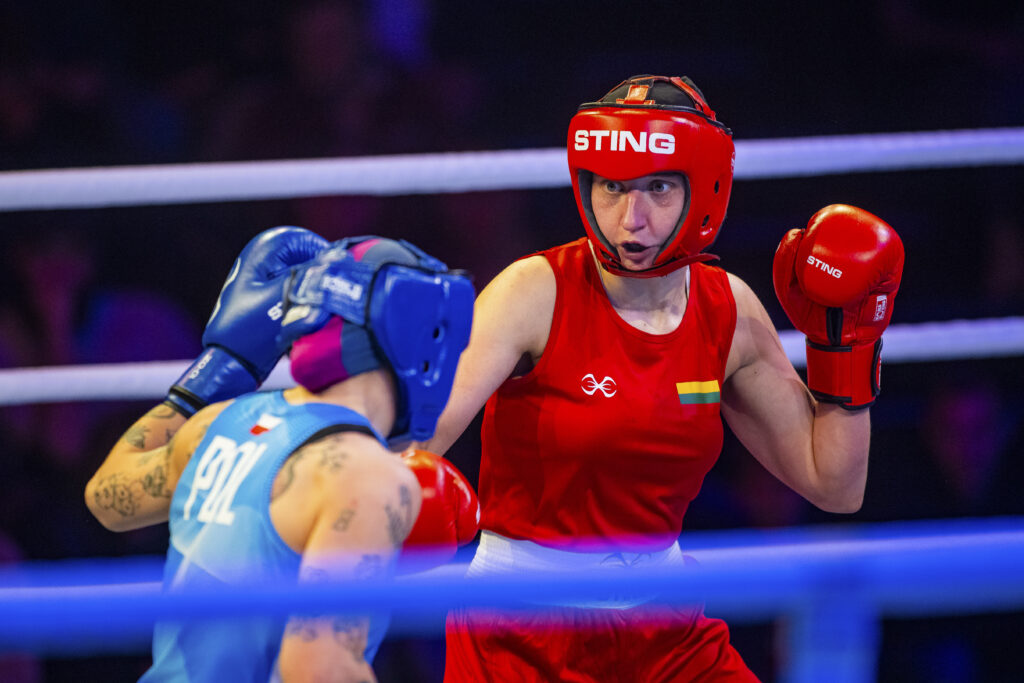 Female boxer in red gear and helmet throws a punch toward the blue-gloved opponent in a ring.