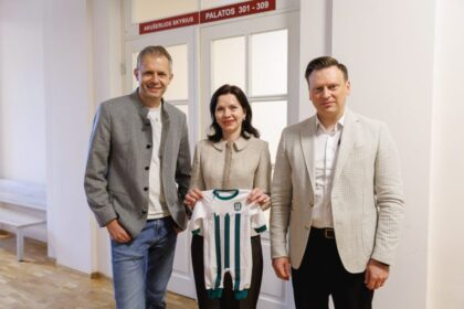 Three adults stand indoors holding a white and green striped baby jersey between them; left man in gray blazer, center woman in beige coat, right man in a light checked blazer.