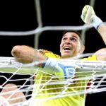 Soccer goalkeeper in a yellow kit reaches up through the goal net with gloved hands, celebrating a save.