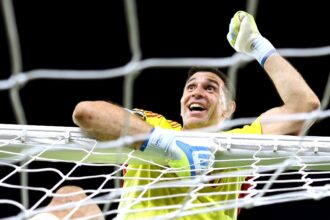 Soccer goalkeeper in a yellow kit reaches up through the goal net with gloved hands, celebrating a save.