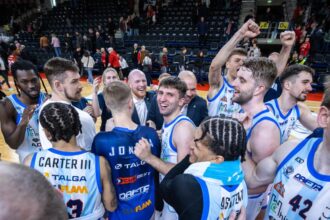 Basketball players in white-and-blue jerseys huddle on the court, celebrating with raised fists and smiles as teammates surround them in a gym arena.