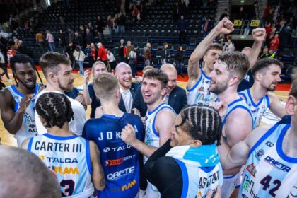 Basketball players in white-and-blue jerseys huddle on the court, celebrating with raised fists and smiles as teammates surround them in a gym arena.