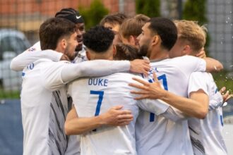 Soccer players in white jerseys form a tight huddle, celebrating on the field.
