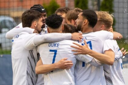 Soccer players in white jerseys form a tight huddle, celebrating on the field.