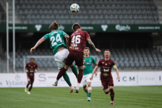 Two opposing soccer players jump to head the ball during a match, one in green (#24 Burba) and one in maroon (#46 Islanddija), with the ball overhead and stadium seats in the background.