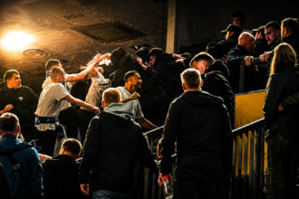Group of men in dark jackets at a stadium railing, cheering and high-fiving amid a crowd under bright lights.