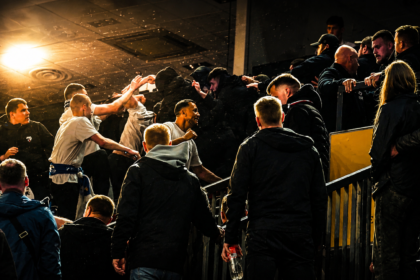 Group of men in dark jackets at a stadium railing, cheering and high-fiving amid a crowd under bright lights.
