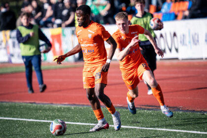 Two soccer players in orange uniforms race for the ball on a green artificial turf field, with spectators in the stands behind them.