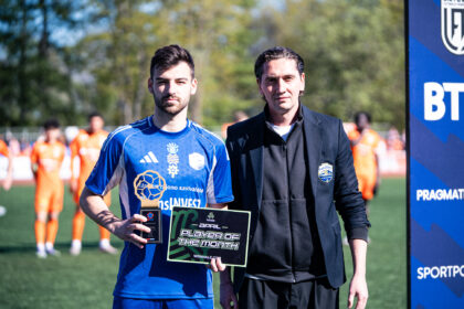 Two men stand on a football field; the left man in a blue jersey holds a 'Player of the Month' plaque.