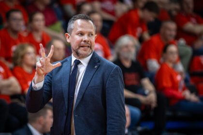 Smiling man in a blue suit making a peace sign with two fingers while speaking at a sports event with a red-clad crowd behind him.
