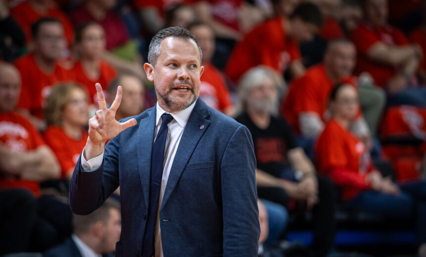 Smiling man in a blue suit making a peace sign with two fingers while speaking at a sports event with a red-clad crowd behind him.