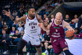 Two basketball players vie for control of the ball during a game, crowd watching from the stands in the background