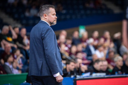 Side profile of a man in a blue blazer standing courtside at a sports event, with a blurred crowd in the background