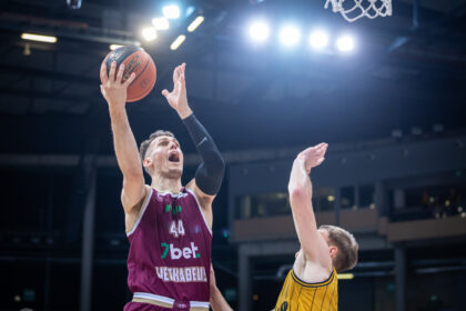 Basketball player in a maroon jersey leaps with the ball toward the basket while a defender reaches up.