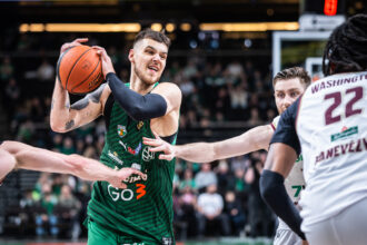 Basketball player in green drives with the ball while a defender in white blocks on a crowded court.