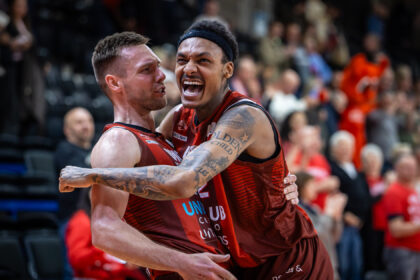 Two basketball players in red jerseys hug in celebration on the court, crowd blurred in the background.