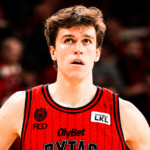 Basketball player in a red jersey, looking upward with a focused expression, crowd blurred in the background