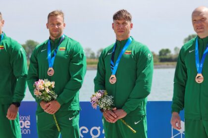 Four male athletes in green tracksuits wear bronze medals with flowers, posing outdoors by the water.