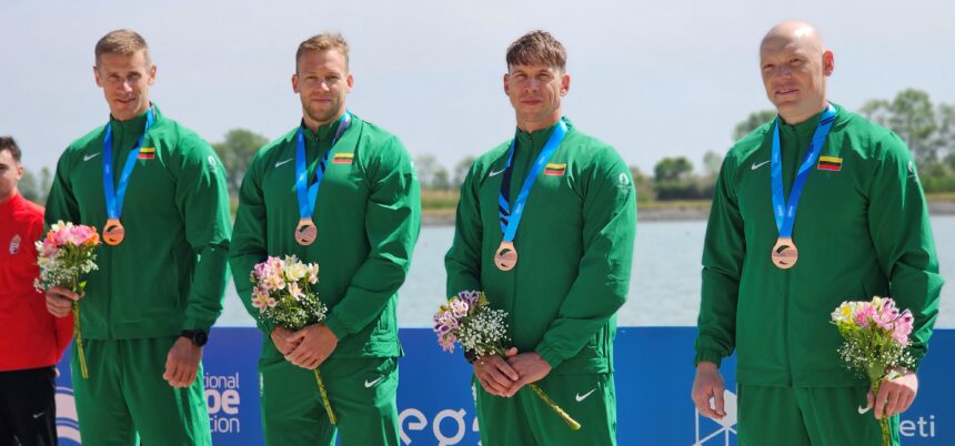 Four male athletes in green tracksuits wear bronze medals with flowers, posing outdoors by the water.