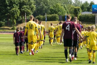 Youth soccer teams in yellow and maroon uniforms walk onto a sunny grass field before a match, hands joined in a line on the way to the field.