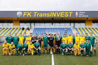 Group of soccer players in green and yellow kits posing on a pitch in front of a stadium stands with blue and yellow seats under a stadium sign reading FK TransINVEST.