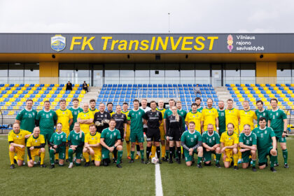 Group of soccer players in green and yellow kits posing on a pitch in front of a stadium stands with blue and yellow seats under a stadium sign reading FK TransINVEST.