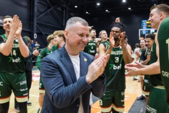 Man in a blue blazer clapping with a basketball team in green uniforms on a gym court during a celebration shift.