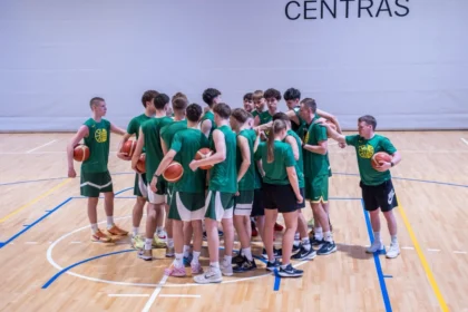 Basketball team in green uniforms forming a huddle on a wooden gym floor, each player holding a ball as a coach leads them.