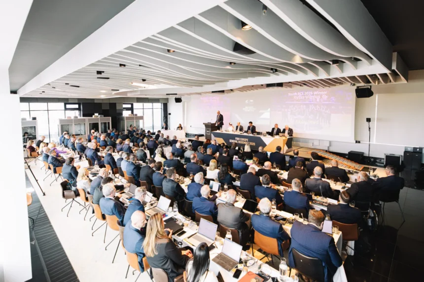 Large conference hall with a panel of speakers at a long table on stage, audience seated at rows of tables listening to the presentation.