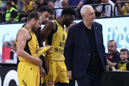 Basketball players in yellow uniforms stand near the sideline as a coach in a dark suit watches; arena spectators in the background.