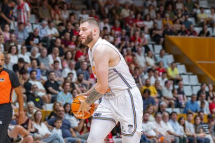 Basketball player in a white jersey grips an orange ball on the court with a crowded arena in the background
