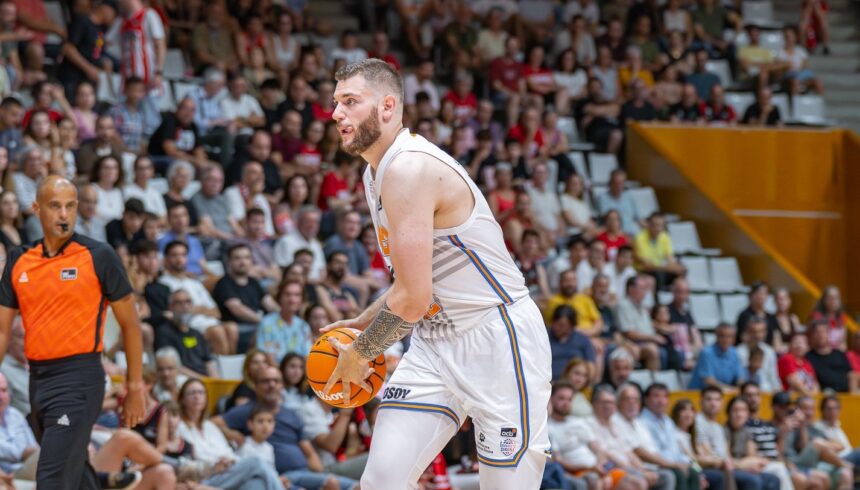 Basketball player in a white jersey grips an orange ball on the court with a crowded arena in the background