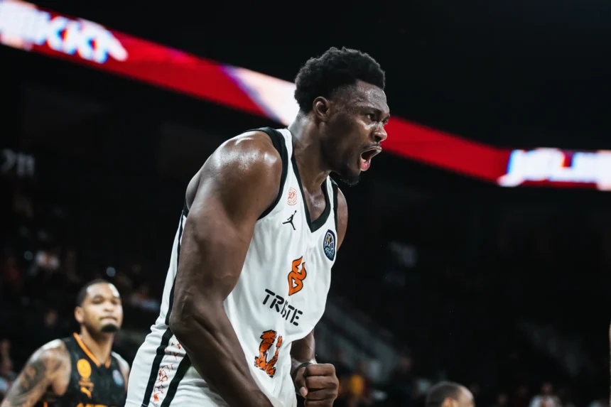 Basketball player in a white jersey shouts passionately during a game, sweat visible on his face and arms with a dark arena behind him.