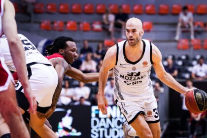 Basketball player in a white uniform dribbles toward the basket as defenders close in on a crowded indoor court.