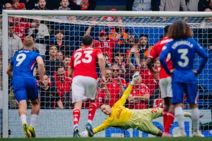 Goalkeeper in yellow dives to block ball at the near post as red attackers press goal; blue and red players surround the area.