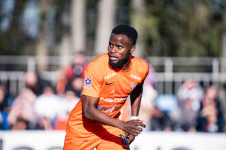 Male soccer player in an orange jersey sprinting on the field with blurred spectators in the background.