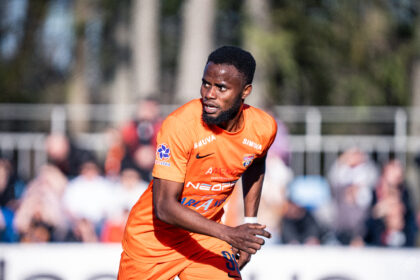 Male soccer player in an orange jersey sprinting on the field with blurred spectators in the background.