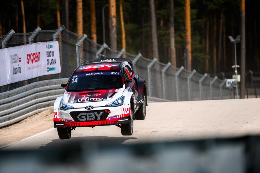 Rally car speeding along a track with forest stands and safety barriers on the side; front of the car shows red, black, and white graphics.